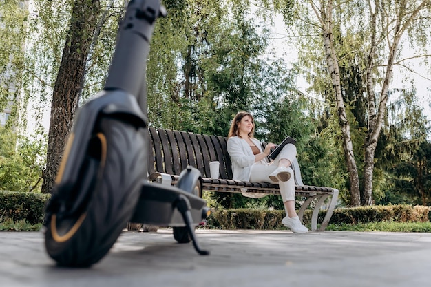 Bonita mujer de negocios con traje blanco sentada en un parque de la ciudad bebe café y trabaja en una tableta digital con un scooter eléctrico en primer plano