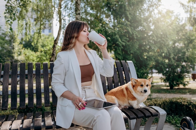 Bonita mujer de negocios con traje blanco sentada en un parque de la ciudad bebiendo café y trabajando en una tableta digital con su perro Welsh Corgi Pembroke