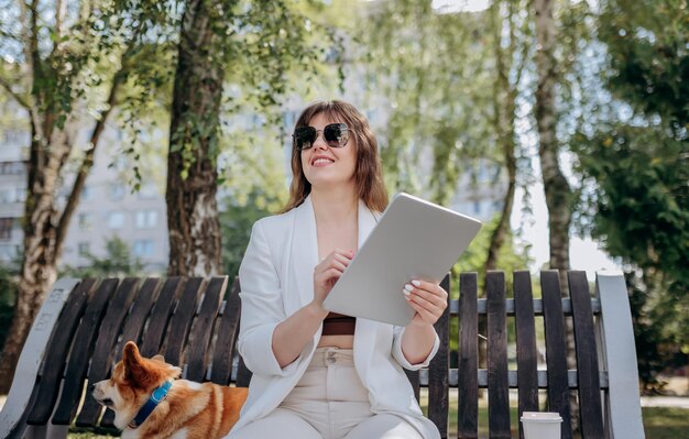 Bonita mujer de negocios con traje blanco sentada en un parque de la ciudad y trabajando en una tableta digital con scooter eléctrico en backgroundxA