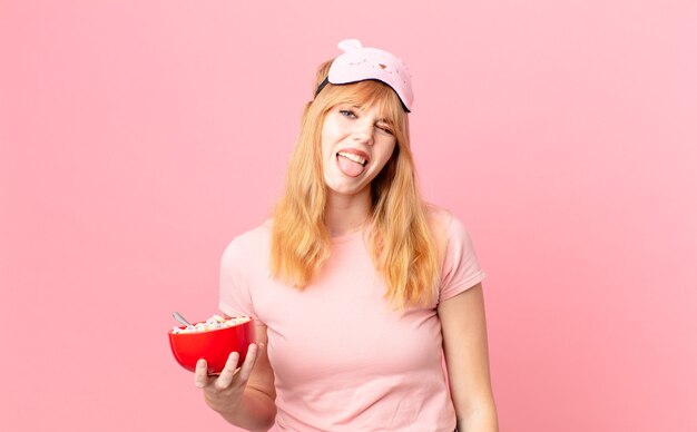 Bonita mujer pelirroja con actitud alegre y rebelde, bromeando y sacando la lengua en pijama y sosteniendo un cuenco de hojuelas