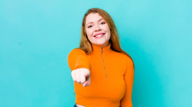 Bonita mujer pelirroja apuntando a la cámara con una sonrisa satisfecha y segura de sí misma eligiéndote