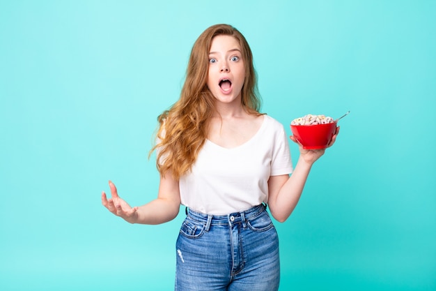 Bonita mujer pelirroja asombrada, conmocionada y asombrada con una increíble sorpresa y sosteniendo un tazón de desayuno