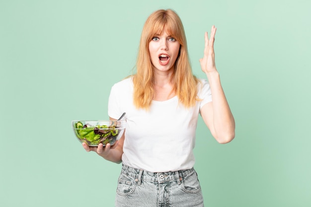 Bonita mujer pelirroja gritando con las manos en el aire y sosteniendo una ensalada. concepto de dieta