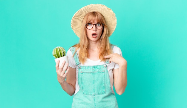 Bonita mujer pelirroja mirando consternado y sorprendido con la boca abierta, apuntando a sí mismo y sosteniendo un cactus en maceta. concepto de granjero