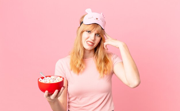 Bonita mujer pelirroja que se siente confundida y perpleja, mostrando que estás loco vistiendo pijamas y sosteniendo un cuenco de hojuelas