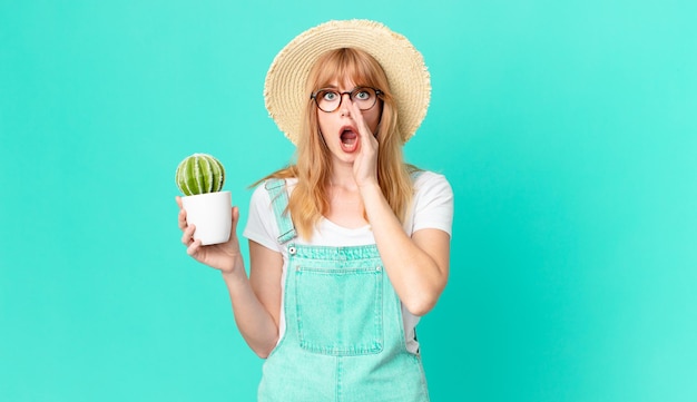 Bonita mujer pelirroja que se siente feliz, dando un gran grito con las manos al lado de la boca y sosteniendo un cactus en maceta. concepto de granjero