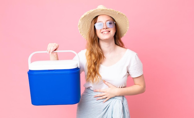 Bonita mujer pelirroja riéndose a carcajadas de una broma hilarante y sosteniendo un refrigerador portátil de picnic