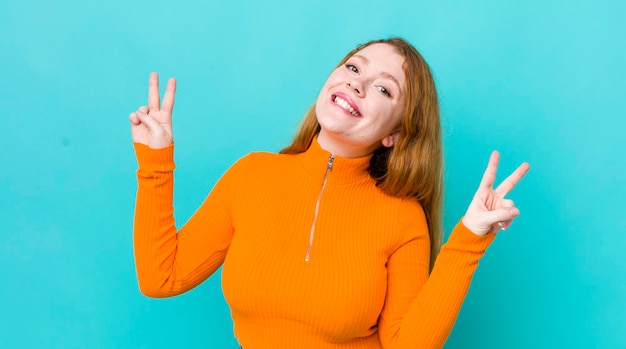 Bonita mujer pelirroja sonriendo y luciendo feliz, amistosa y satisfecha, gesticulando victoria o paz con ambas manos