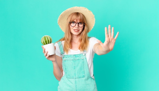 Bonita mujer pelirroja sonriendo y mirando amigable, mostrando el número cinco y sosteniendo un cactus en maceta. concepto de granjero