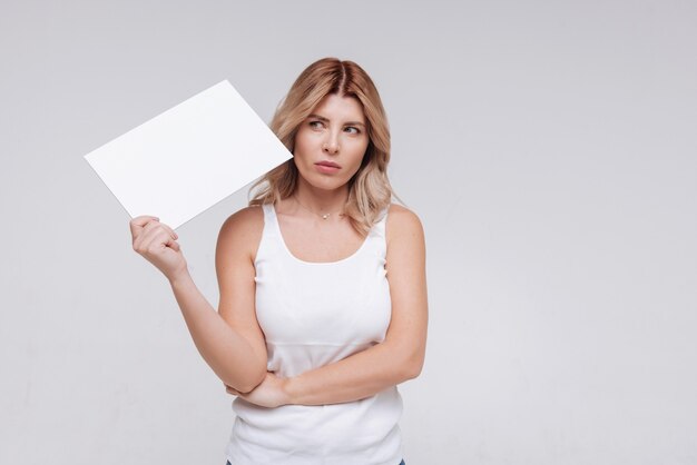 Bonita mujer preguntándose acerca de algo mientras sostiene una hoja de papel en blanco