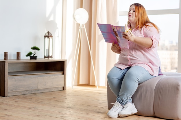 Bonita mujer regordeta comiendo un sabroso plátano mientras lee una revista