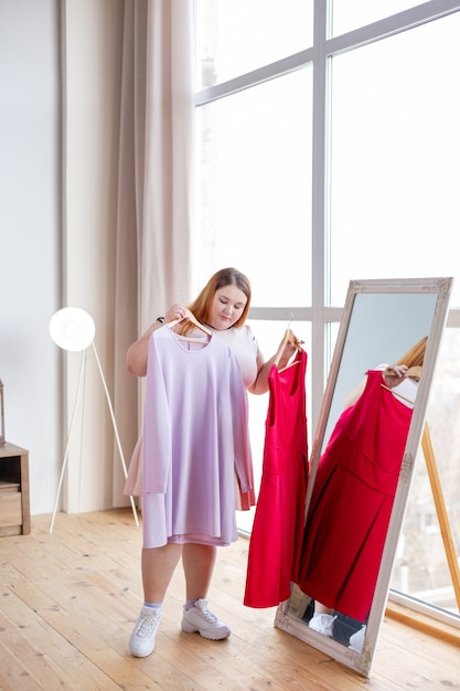 Bonita mujer regordeta de pie frente al espejo mientras sostiene dos perchas con vestidos