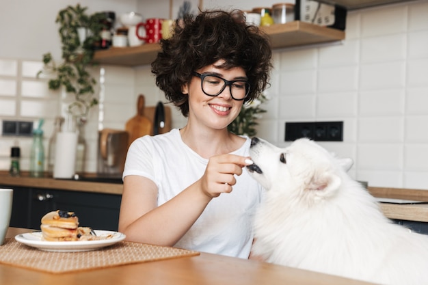 Bonita mujer rizada sentada en la cocina comer pastel y alimentar a su lindo perro blanco