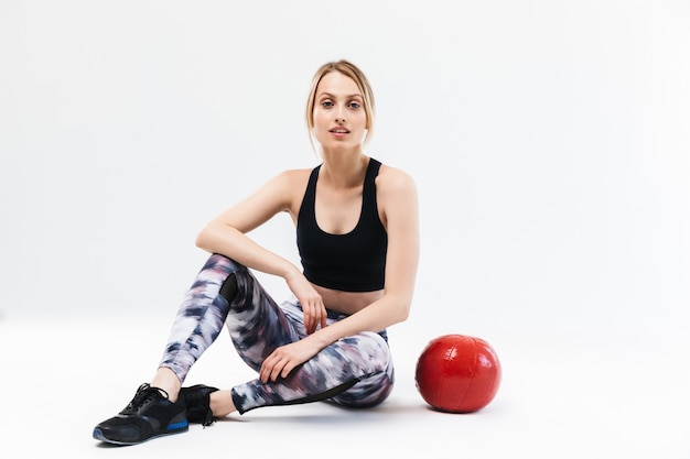 Bonita mujer rubia de 20 años vestida con ropa deportiva haciendo ejercicio y haciendo ejercicios con pelota de fitness durante aeróbic aislado sobre una pared blanca