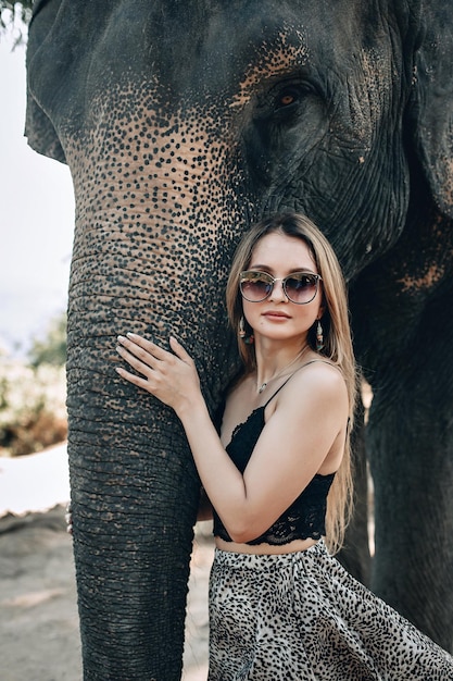 Bonita mujer rubia con elegantes gafas de sol y un vestido con estampado de leopardo abraza suavemente la trompa de elefante; concepto de amistad.