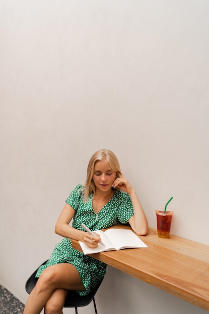 Bonita mujer rubia escribiendo en un cuaderno con vestido de verano sentada en un café moderno con paredes blancas