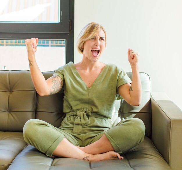 Bonita mujer rubia gritando triunfalmente, con aspecto de ganador emocionado, feliz y sorprendido, celebrando