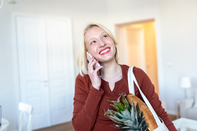 Bonita mujer rubia que llega a casa llevando comestibles en una bolsa de mano ecológica usando su teléfono móvil inteligente