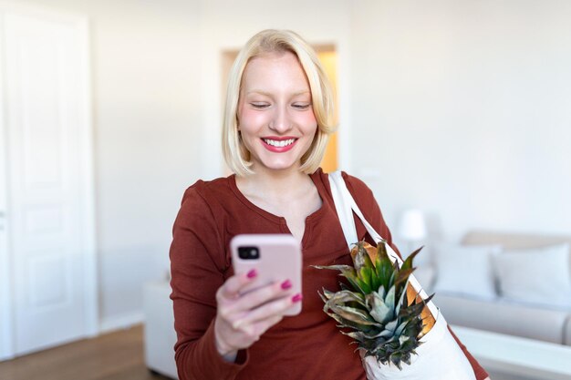 Bonita mujer rubia que llega a casa llevando comestibles en una bolsa de mano ecológica usando su teléfono móvil inteligente