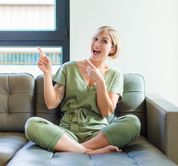 Bonita mujer rubia que se siente alegre y sorprendida, sonriendo con una expresión de asombro y apuntando hacia un lado