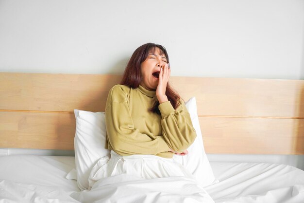 Bonita mulher asiática se estendendo e bocejando enquanto está sentada em sua cama no quarto brilhante