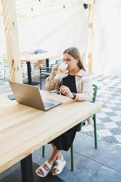 Bonita mulher europeia sentada na mesa de café ao ar livre com laptop e xícara de café mulher sorridente em óculos desfrutando de teletrabalho no café ou estudando online