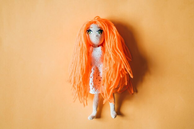 Bonita muñeca hecha a mano de punto con pelo largo naranja, con vestido blanco. Juguete para niños lindo aislado sobre fondo melocotón. Crochet para niños. Hecho a mano, niños, concepto de pasatiempos. Vista superior.