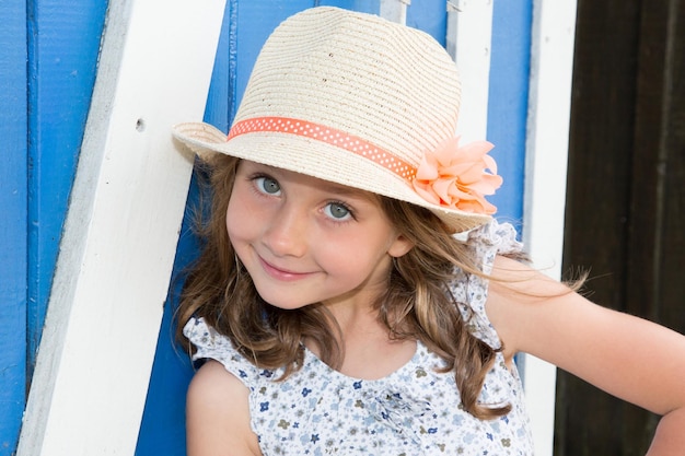 Bonita niña de belleza con vestido de flores y sombrero de paja sonriendo con fondo de cabaña blanca y azul de madera