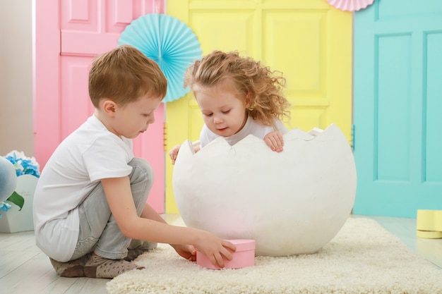 Bonita niña rubia rizada está sentada en un huevo blanco y adorable retrato de niño rubio en blanco