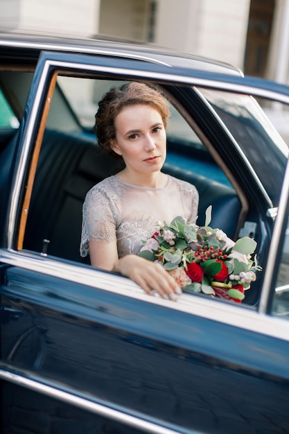 Bonita novia en vestido de novia de lujo sentado dentro del coche retro y con ramo de novia