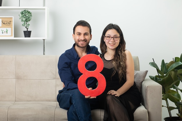 Bonita pareja sonriente sosteniendo la figura roja de ocho sentados en el sofá en la sala de estar en el día internacional de la mujer de marzo