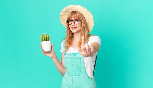 bonita pelirroja sonriendo con orgullo y confianza haciendo el número uno y sosteniendo un cactus en maceta. concepto de granjero