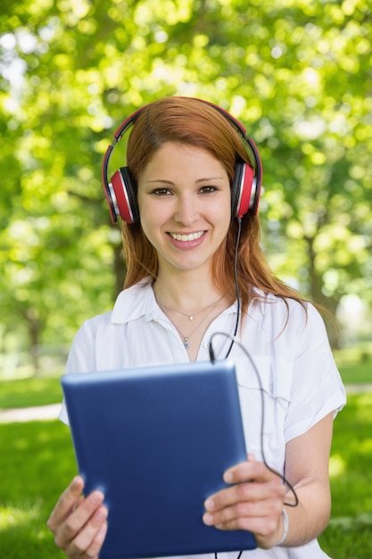 Bonita pelirroja usando su tablet pc mientras escucha música en el parque