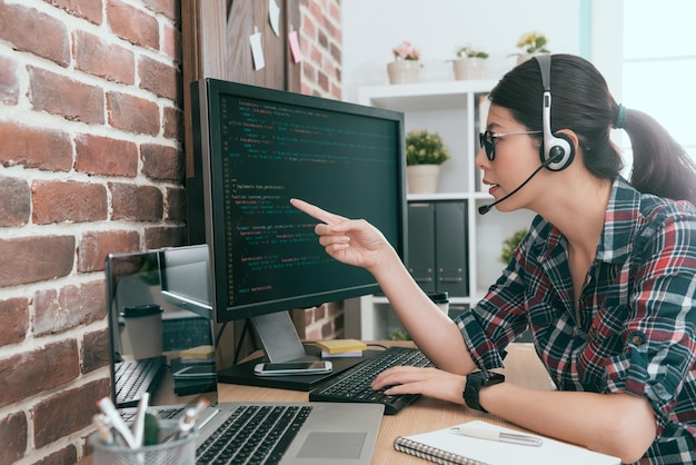 bonita programadora de belleza mujer con micrófono de auriculares hablando con un colega discutiendo el problema de reparación del sistema remoto y mirando la codificación de datos de monitoreo de la pantalla de la computadora.