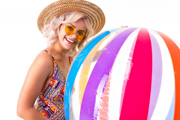 Bonita rubia mujer caucásica se encuentra en traje de baño con pelota de goma grande playa colorida y sonrisas aisladas