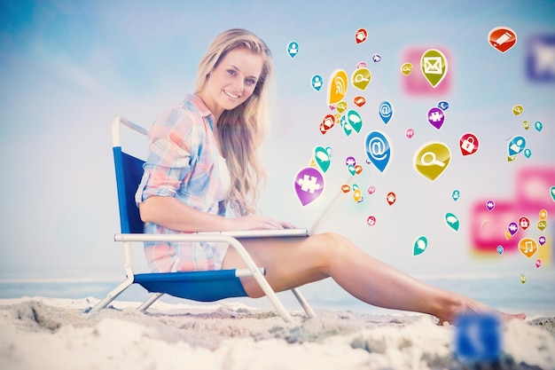 Bonita rubia sentada en la playa usando su computadora portátil con aplicaciones informáticas coloridas