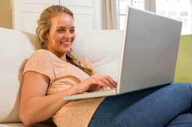 Bonita rubia usando laptop sentado en el sofá de la sala de estar