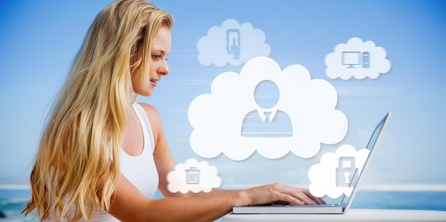 Bonita rubia usando su computadora portátil en la playa contra nubes e íconos