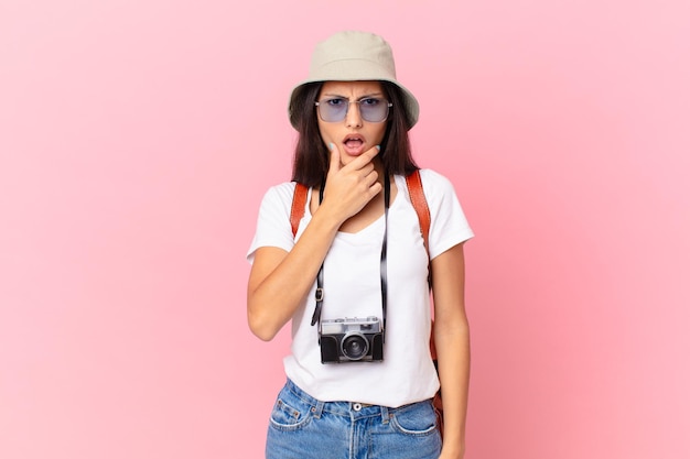 Bonita turista hispana con la boca y los ojos bien abiertos y la mano en la barbilla con una cámara de fotos y un sombrero