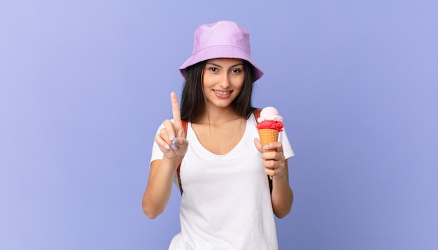 Bonita turista hispana sonriendo con orgullo y confianza haciendo el número uno y sosteniendo un helado