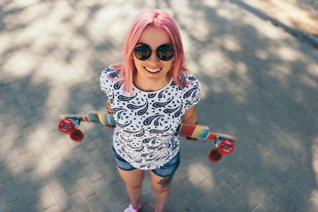 Bonita vista superior de una atractiva mujer joven con cabello rosado y longboard en la calle con gafas de sol mirando hacia arriba Hermosa sonrisa chica caminando con longboard en la calle Estilo de vida