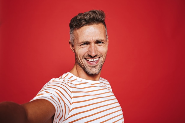 Bonitão, 30 anos, com camiseta listrada, sorrindo enquanto tira selfie foto isolada no vermelho