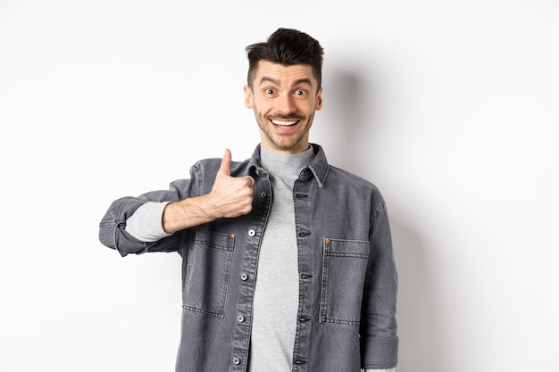 Bonitão animado mostrando o polegar para cima e sorrindo, como coisa boa, elogia a escolha incrível, parecendo satisfeito e feliz, em pé sobre um fundo branco.