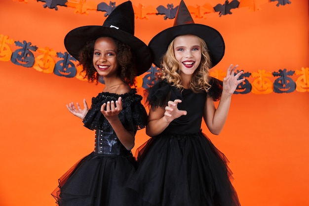 Bonitas chicas multinacionales en disfraces de halloween negros sonriendo y burlándose aislado sobre la pared de calabaza naranja