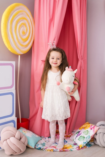 Bonitinha em um vestido inteligente no fundo de um quarto de criança linda. unicórnio de brinquedo em suas mãos.