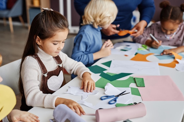 Bonito aluno diligente da creche criando brinquedo de papel enquanto está sentado à mesa entre seu colega de classe