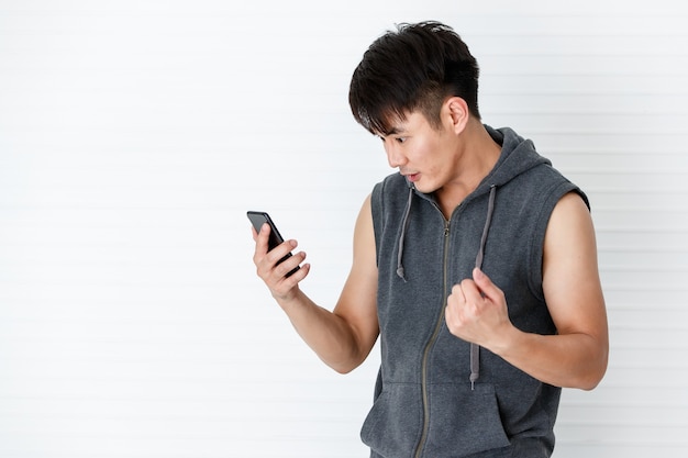 Bonito asiático apto sorridente homem segurando a mão smartphone gesto feliz vestindo sportswear cinza t-shirt sem mangas em fundo branco.