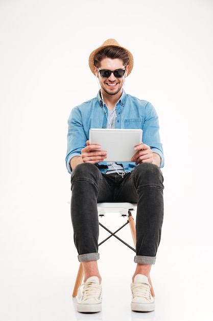 Bonito atraente sorridente jovem feliz usando tablet e sentado na cadeira isolado