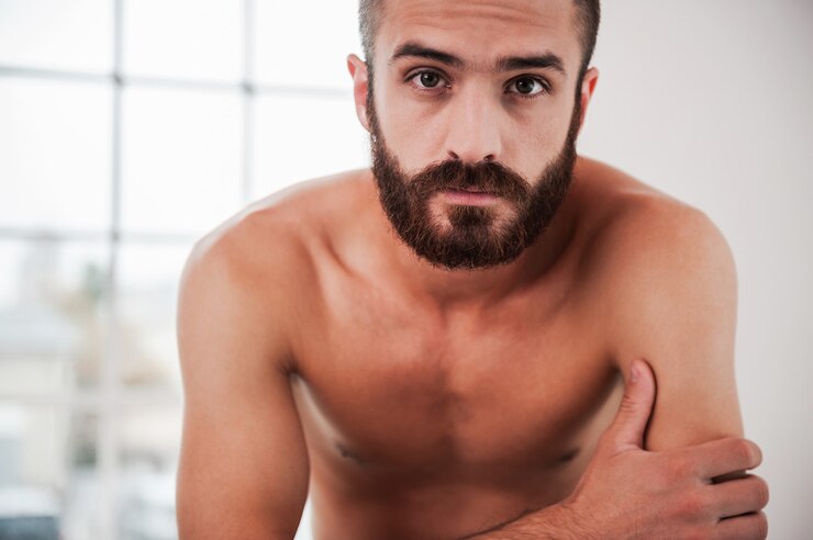 Foto bonito barbudo. jovem confiante, barbudo e sem camisa, olhando para a câmera enquanto está sentado dentro de casa