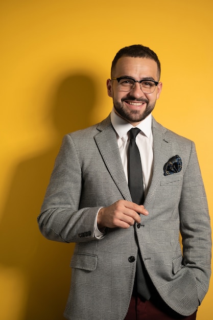 Bonito barbudo sorridente homem elegante usando óculos com roupa formal com uma mão levantada em pé isolado no fundo amarelo.